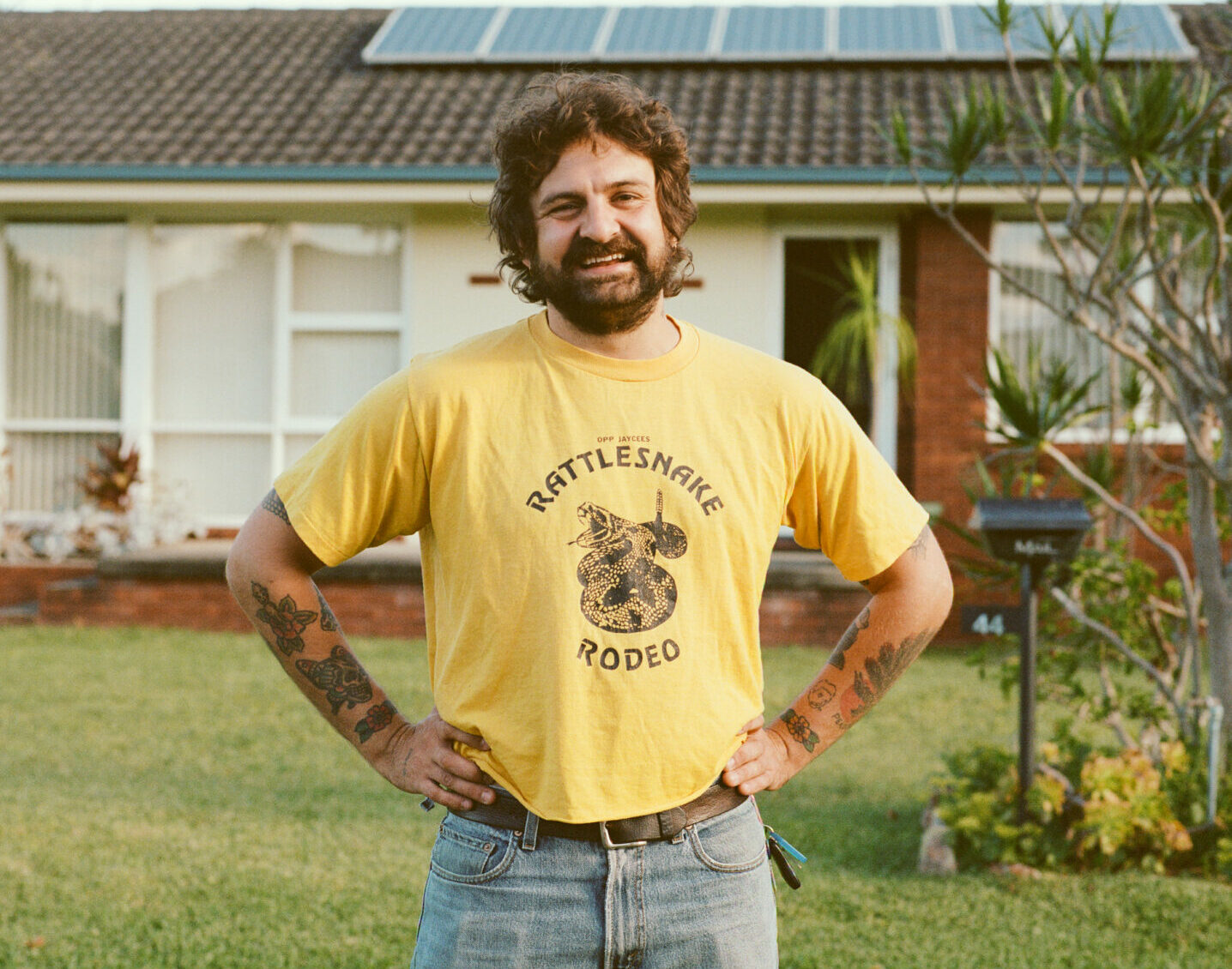 A man with brown hair and a full beard, wearing a yellow t-shirt and standing with his hands on his hips
