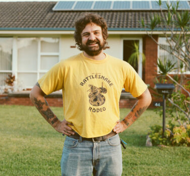 A man with brown hair and a full beard, wearing a yellow t-shirt and standing with his hands on his hips