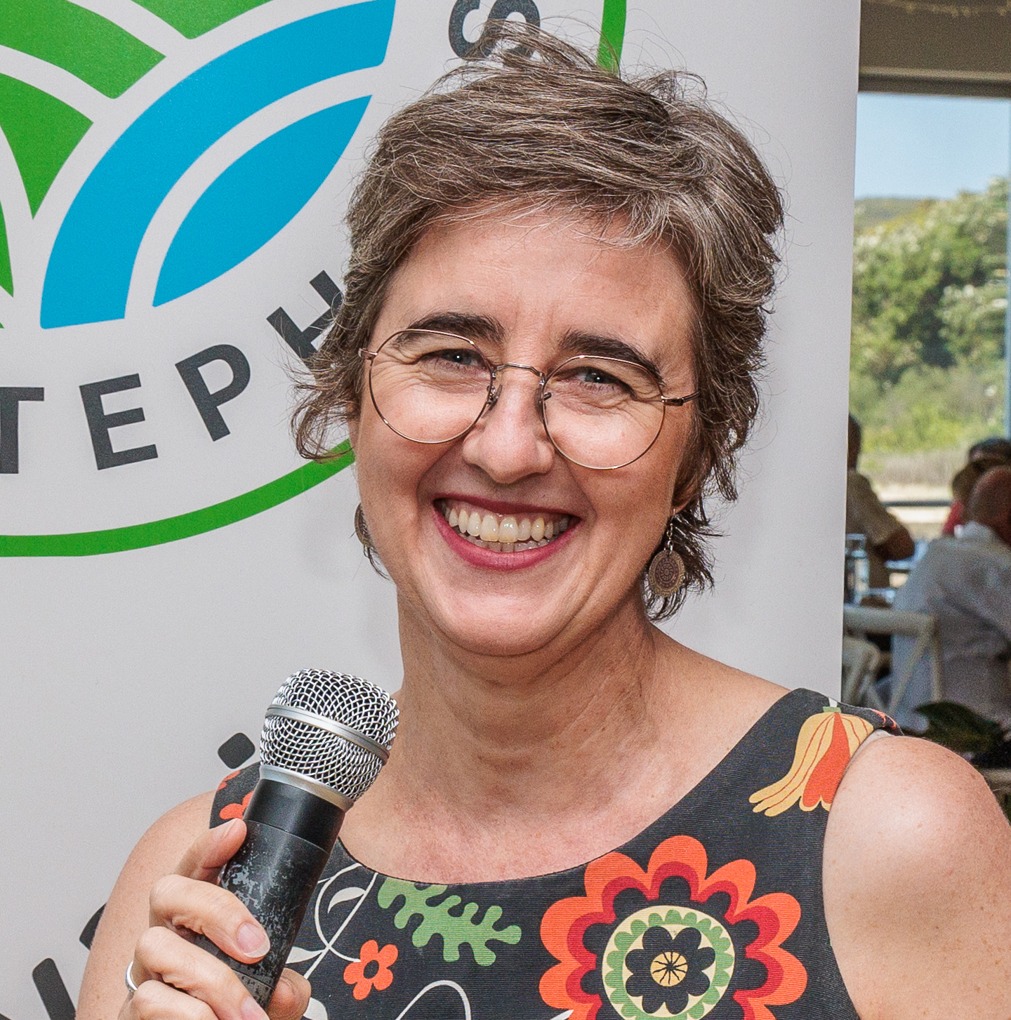 A woman with short gray-brown hair and glasses, holding a microphone and smiling.