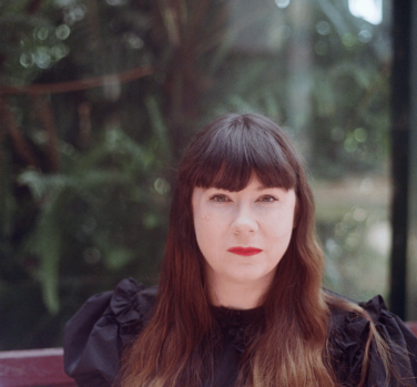 A woman with long brown hair and a fringe, wearing red lipstick and looking directly at the camera