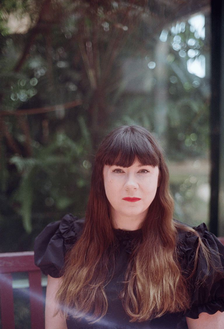 A woman with long brown hair and a fringe, wearing red lipstick and looking directly at the camera