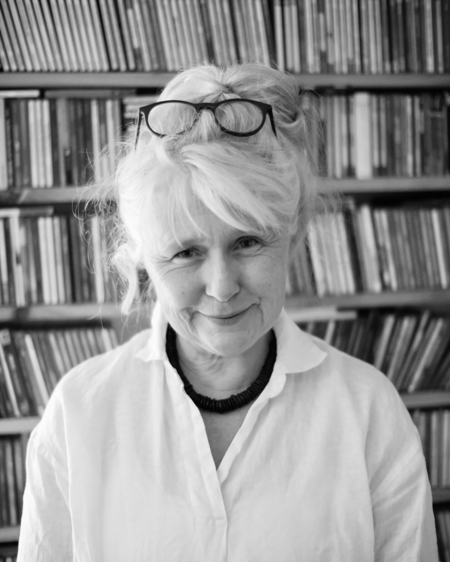 A black and white photograph of a woman with glasses on top of her bun, standing in front of a wall of books