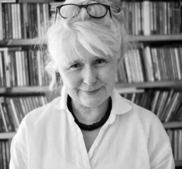 A black and white photograph of a woman with glasses on top of her bun, standing in front of a wall of books