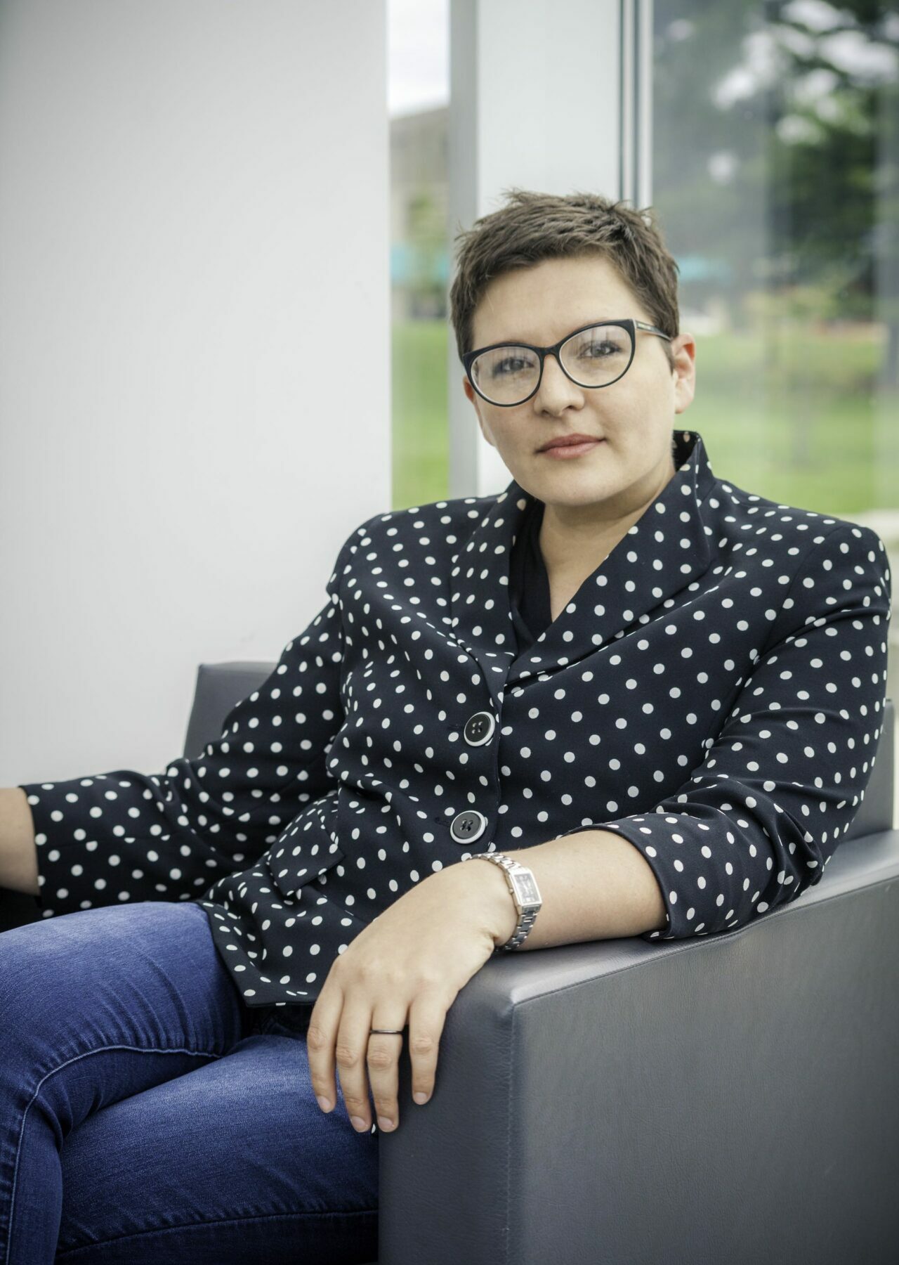 A photograph of a woman with short brown hair and glasses, sitting comfortably in a chair