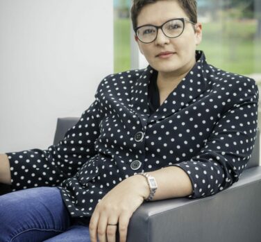 A photograph of a woman with short brown hair and glasses, sitting comfortably in a chair