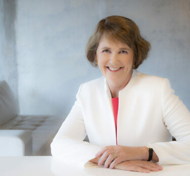 A woman with short brown hair in a white suit leaning on a desk and smiling