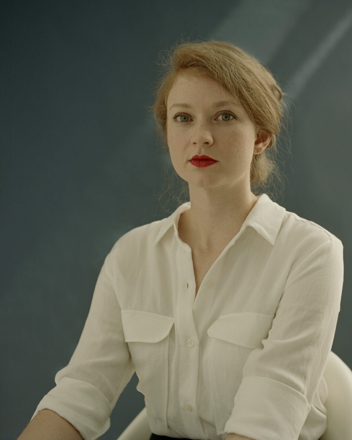 A strawberry-blonde woman with red lipstick and a white shirt sits up and looks directly at the camera