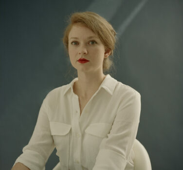 A strawberry-blonde woman with red lipstick and a white shirt sits up and looks directly at the camera