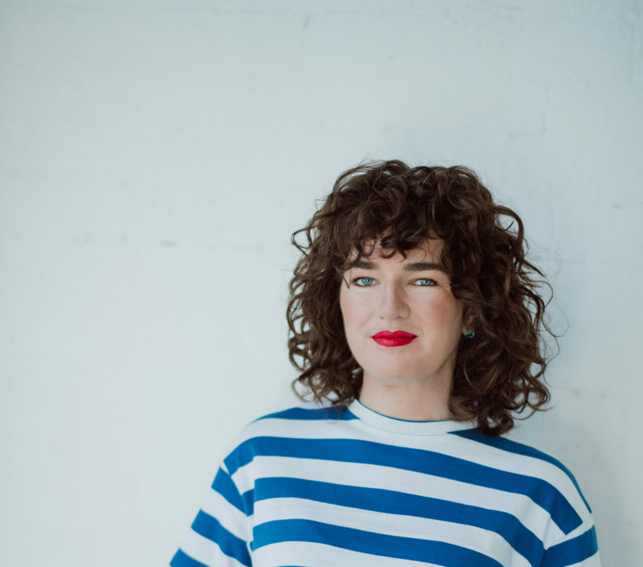 A woman with brown curly hair and bright red lipstick, wearing a striped blue and white shirt, leans against a white background
