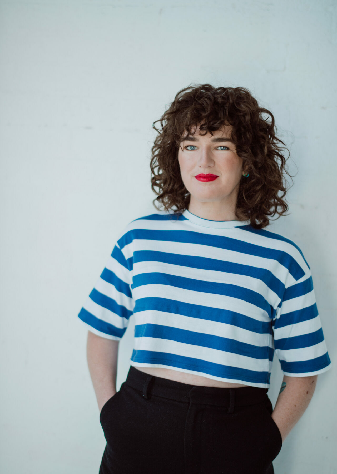 A woman with brown curly hair and bright red lipstick, wearing a striped blue and white shirt, leans against a white background