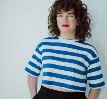 A woman with brown curly hair and bright red lipstick, wearing a striped blue and white shirt, leans against a white background