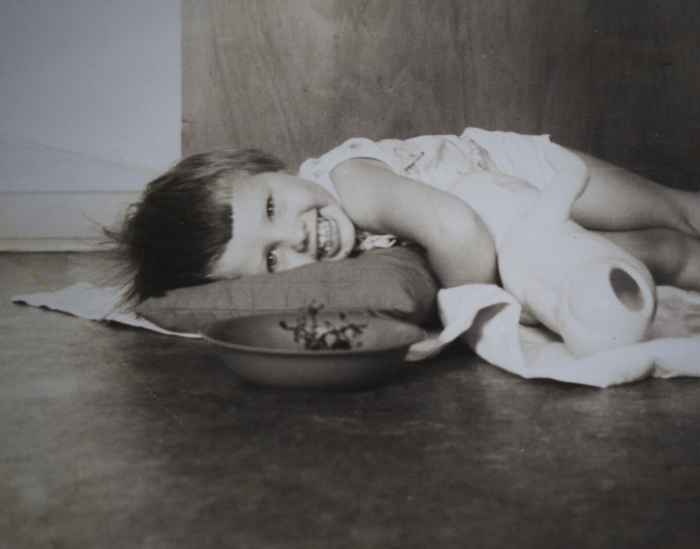 A black and white photograph of a smiling child lying on a pillow on the floor behind a bowl