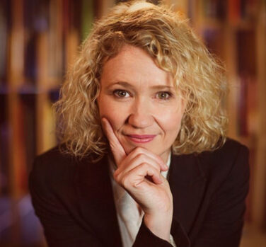 A curly-haired blonde woman leans on her hand and smiles, looking directly at the camera