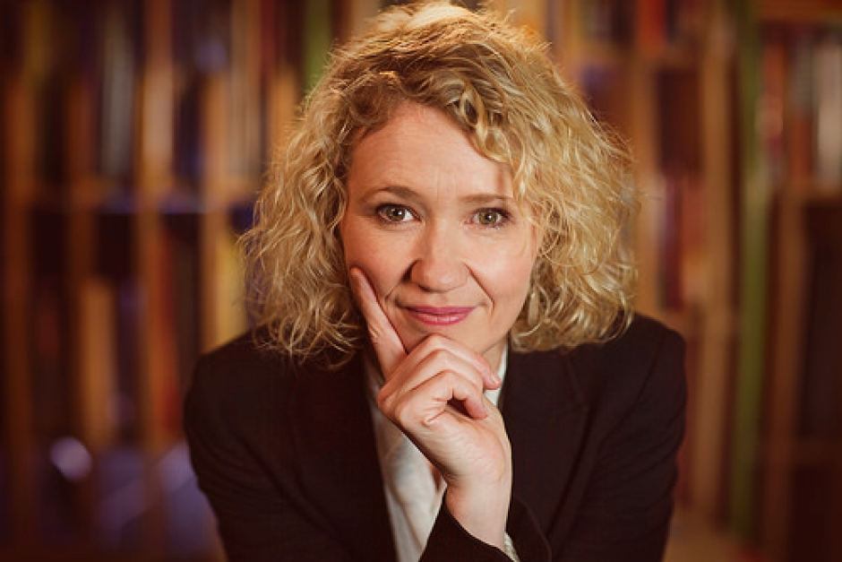 A curly-haired blonde woman leans on her hand and smiles, looking directly at the camera