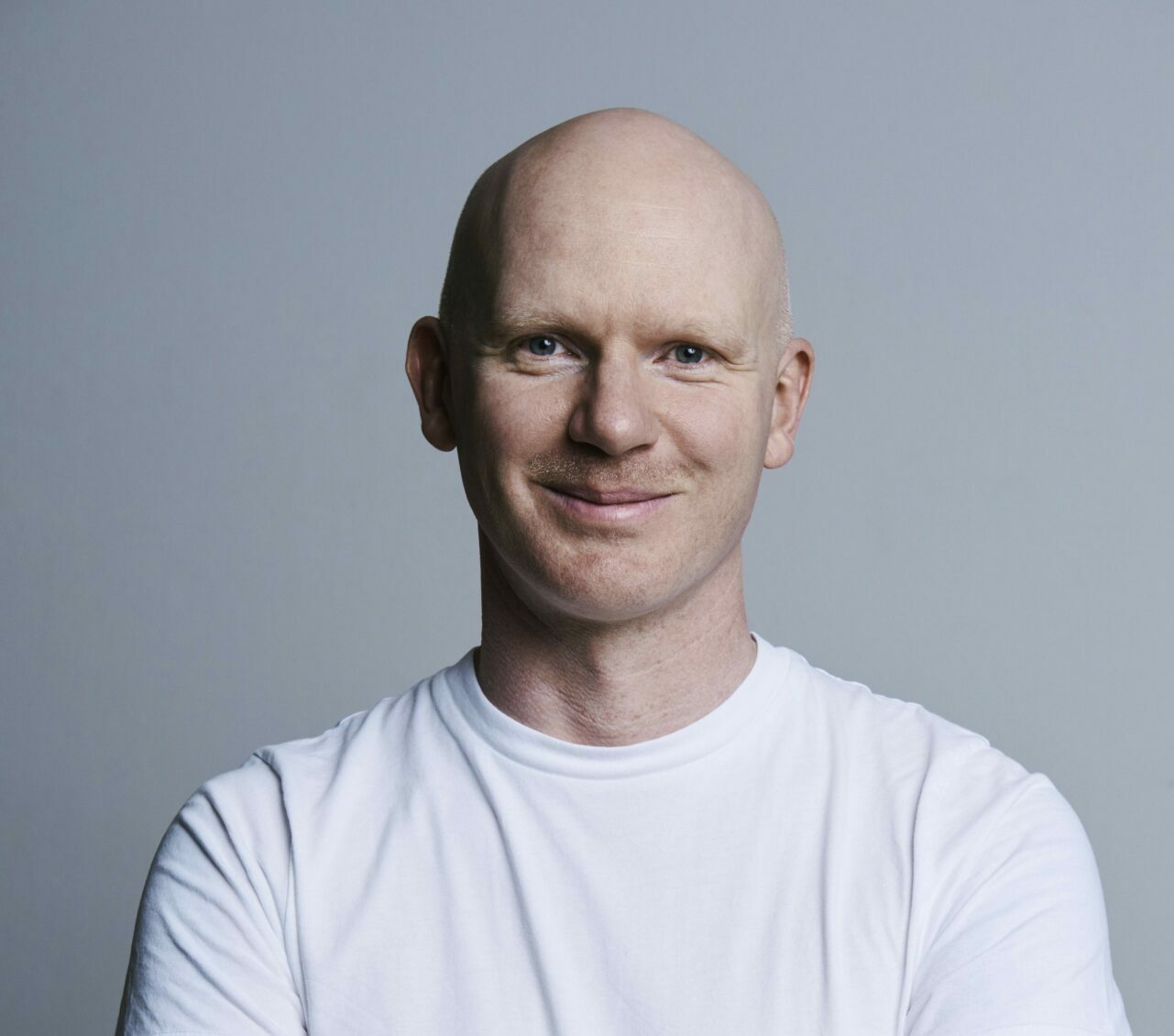 A man in a white t-shirt with a shaved/bald head stands arms folded looking directly at the camera and smiles