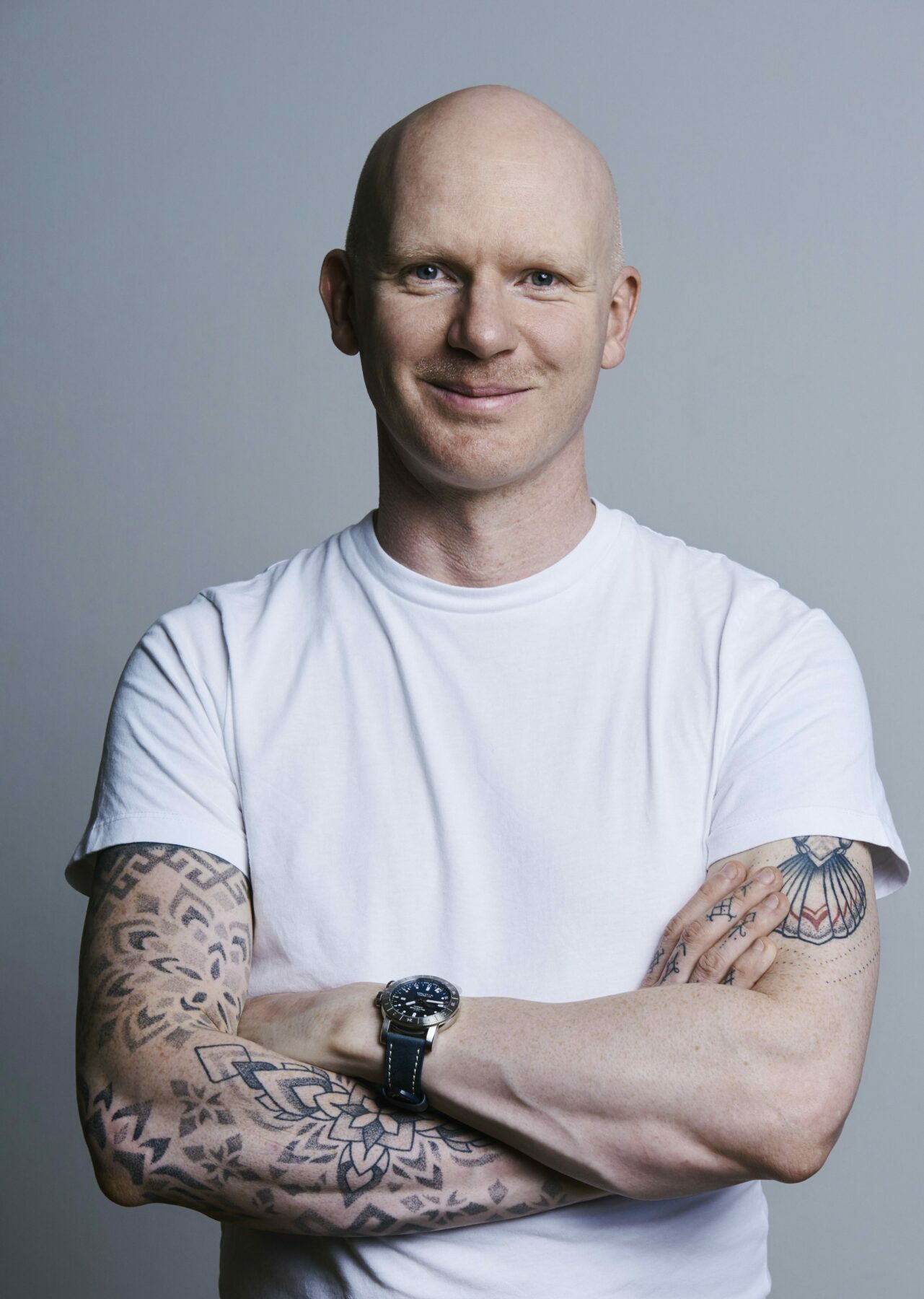 A man in a white t-shirt with a shaved/bald head stands arms folded looking directly at the camera and smiles