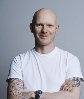 A man in a white t-shirt with a shaved/bald head stands arms folded looking directly at the camera and smiles