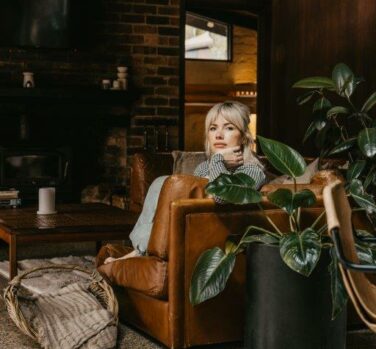 A photograph of a blonde woman sitting on a sofa in a lounge room, her body obscured by the side of the sofa and a pot plant