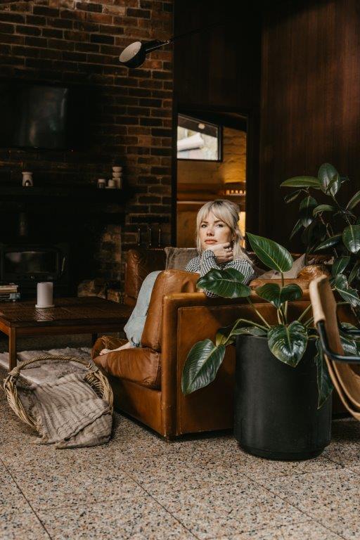A photograph of a blonde woman sitting on a sofa in a lounge room, her body obscured by the side of the sofa and a pot plant