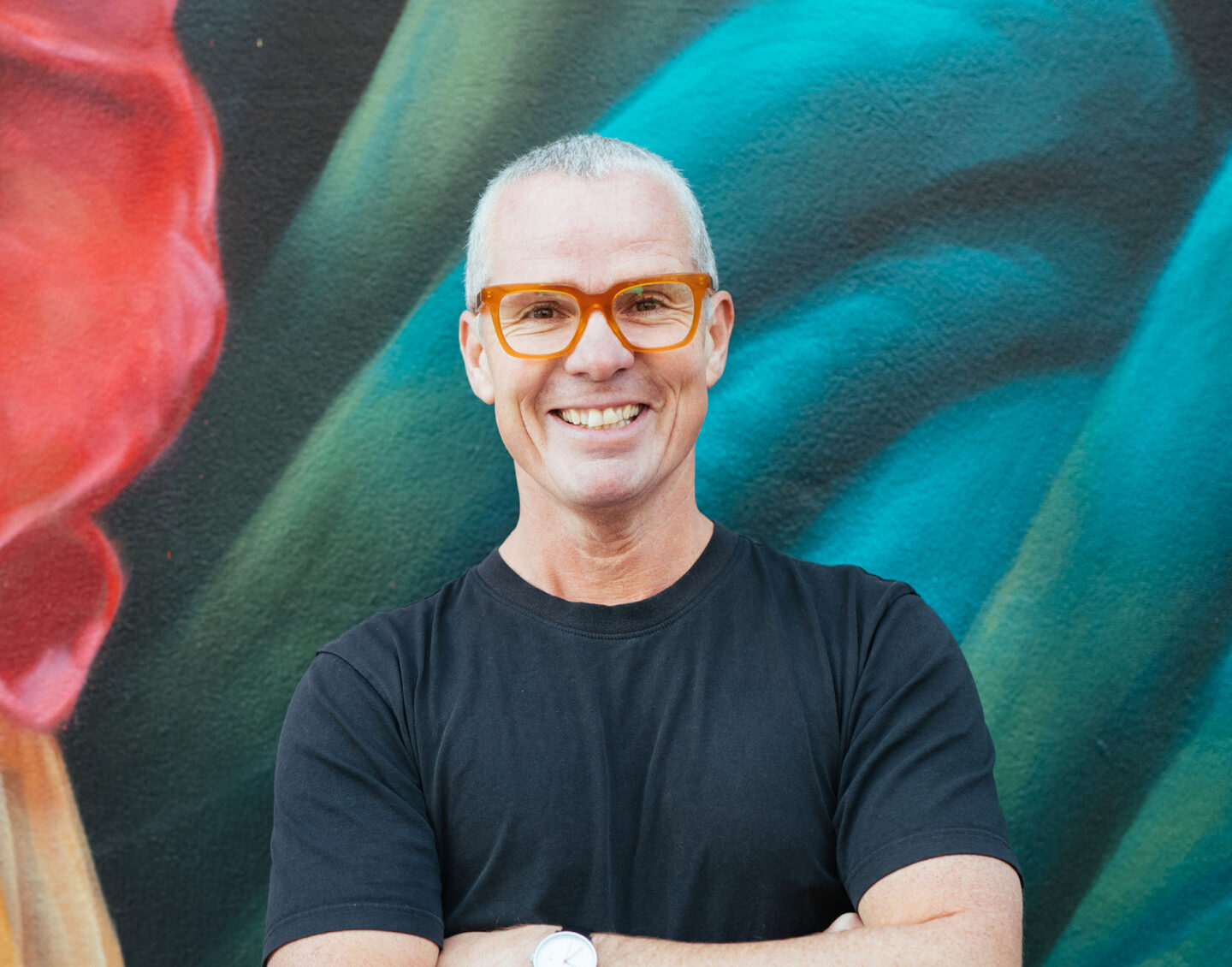 A man with short gray hair and red-rimmed glasses stands arms-folded and smiling in front of a colourful artwork.