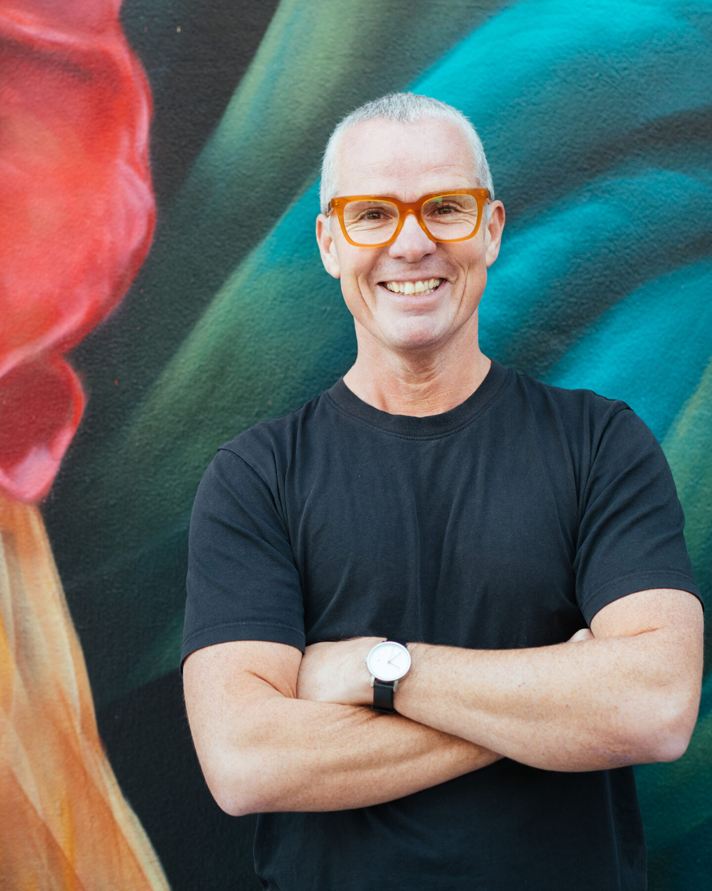 A man with short gray hair and red-rimmed glasses stands arms-folded and smiling in front of a colourful artwork.