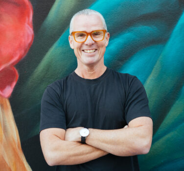 A man with short gray hair and red-rimmed glasses stands arms-folded and smiling in front of a colourful artwork.