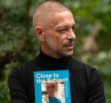 A brown-skinned man with a shaved head in a black long-sleeved shirt looks off to the side, while holding a copy of his book.