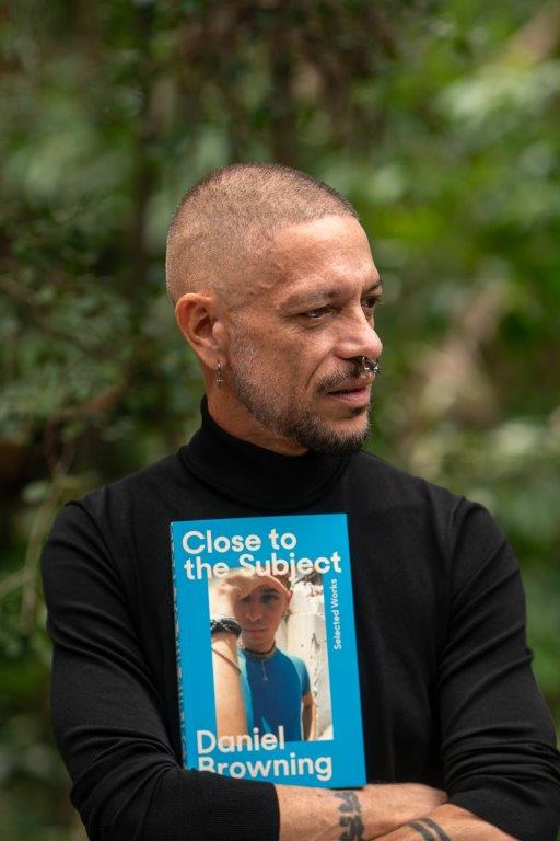 A brown-skinned man with a shaved head in a black long-sleeved shirt looks off to the side, while holding a copy of his book.