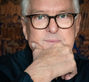 A white man with short grey hair and dark-rimmed glasses, rubbing his chin