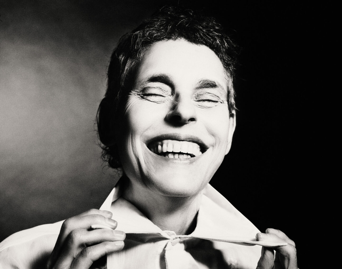 A black and white headshot of a laughing woman with short dark hair, pulling on her collar with both hands