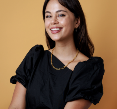 A photograph of a smiling woman with long dark hair and a gold necklace, leaning back at an angle