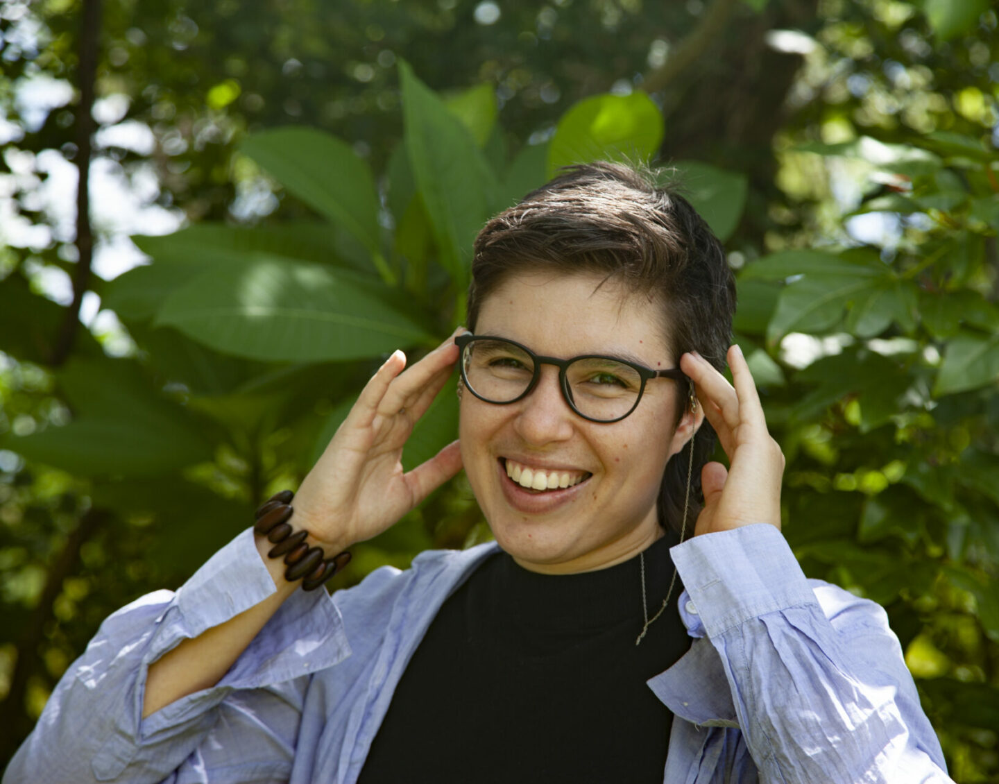 A person with short brown hair smiles and touches both hands to their dark-rimmed glasses, against a backdrop of green plants