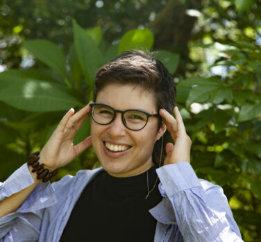 A person with short brown hair smiles and touches both hands to their dark-rimmed glasses, against a backdrop of green plants