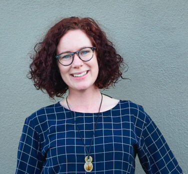 A head and shoulders photograph of a woman with curly dark auburn hair and glasses wearing a blue checked dress