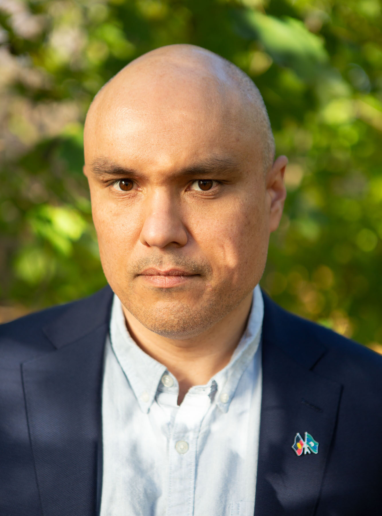 A head-and-shoulders photograph of a bald man with light brown skin looking directly at the camera without smiling