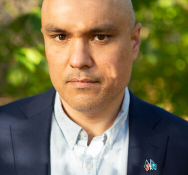 A head-and-shoulders photograph of a bald man with light brown skin looking directly at the camera without smiling