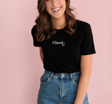 A woman with wavy brown hair smiles at the camera with one hand behind her back. She is wearing a black t-shirt that reads 'cheek'