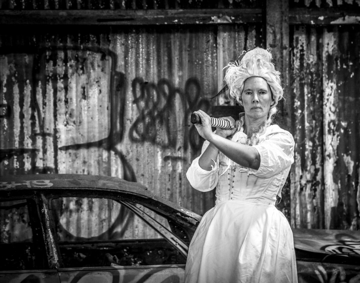 A black and white photograph of a woman in white period clothing, holding something with a long handle over her shoulder. She stands in front of a car and wall of corrugated metal, both covered in graffiti tags