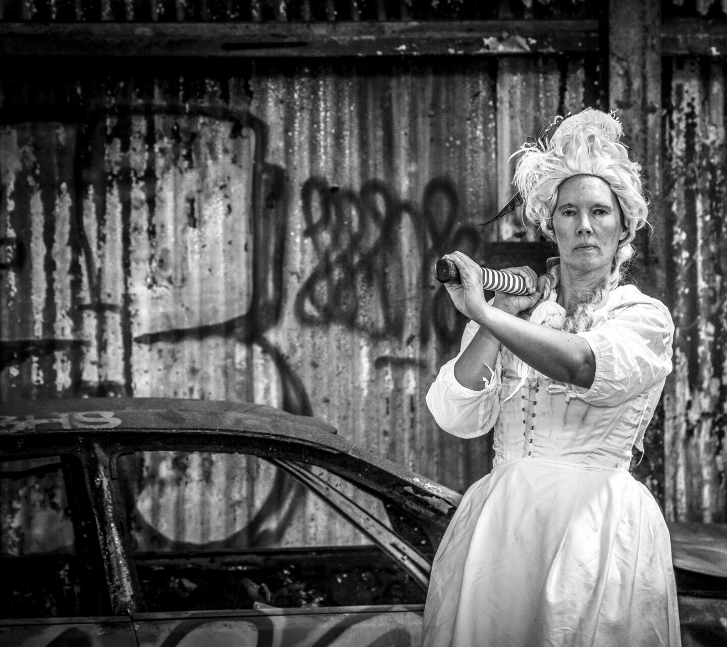 A black and white photograph of a woman in white period clothing, holding something with a long handle over her shoulder. She stands in front of a car and wall of corrugated metal, both covered in graffiti tags