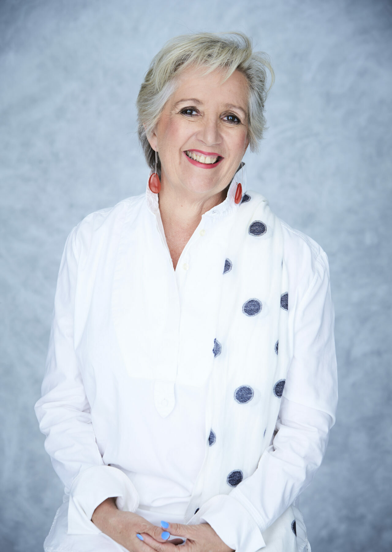 A smiling middle-aged woman with brown eyes and short grey hair smiles with her hands clasped in front of her