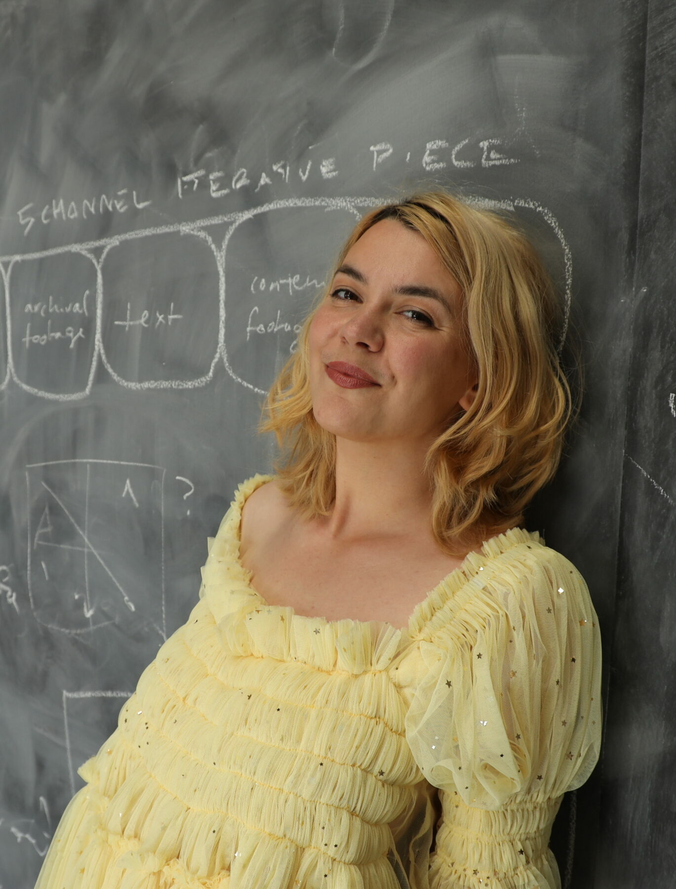 A person with shoulder-length blonde hair and red lipstick leans against a blackboard and smiles closed-lipped