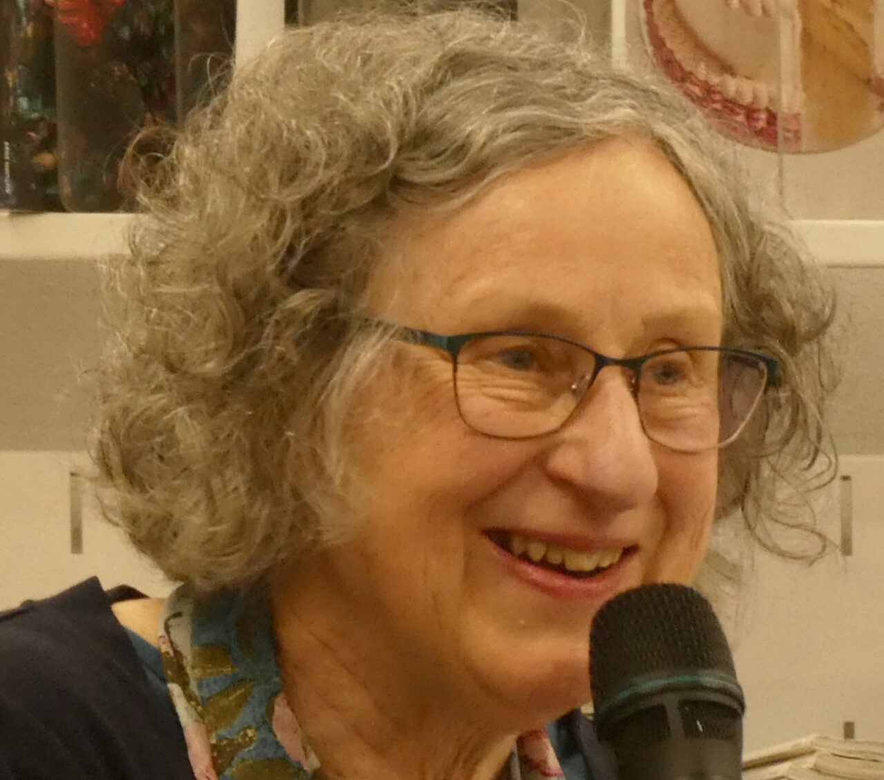 A grey-haired woman with glasses and a floral scarf leans forward an speaks into a handheld microphone
