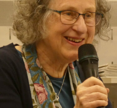 A grey-haired woman with glasses and a floral scarf leans forward an speaks into a handheld microphone