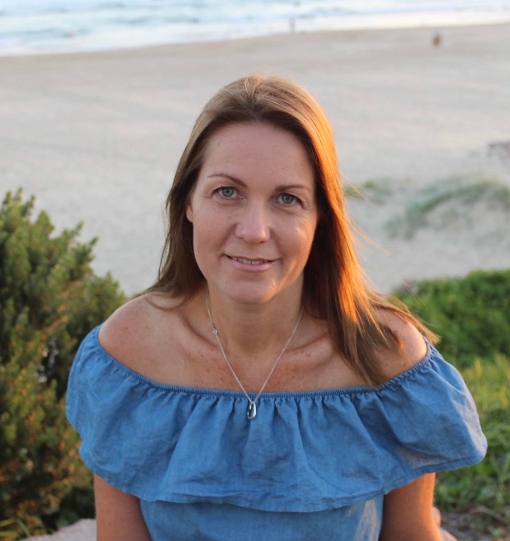 A woman with straight brown hair and a blue top looks at the camera and smiles, with the beach behind her