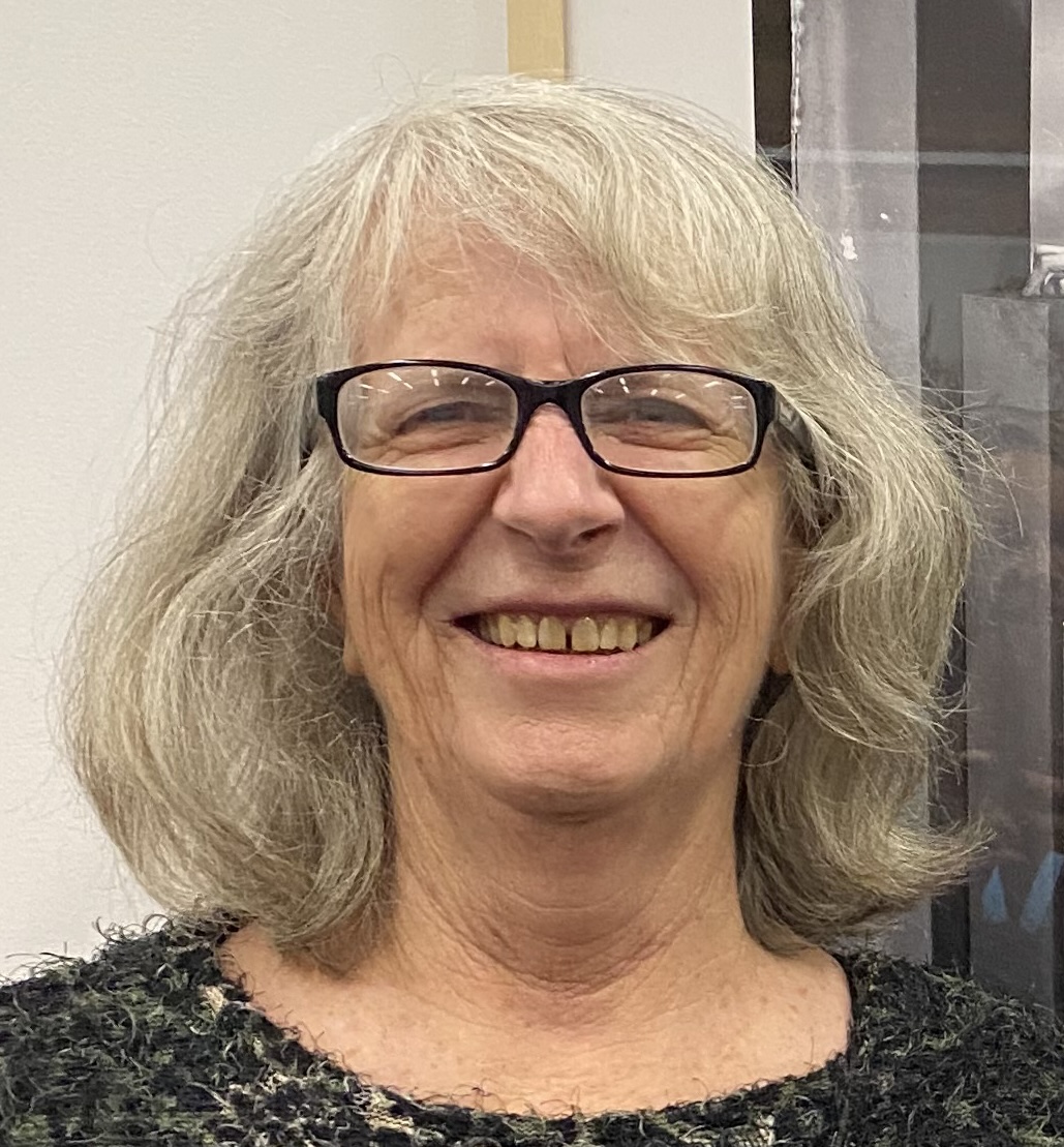A middle-aged woman with wavy shoulder-length gray hair and black rectangular-framed glasses smiles at the camera