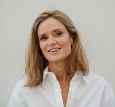 A white woman with shoulder-length dark blonde hair and a white blouse smiles at the camera