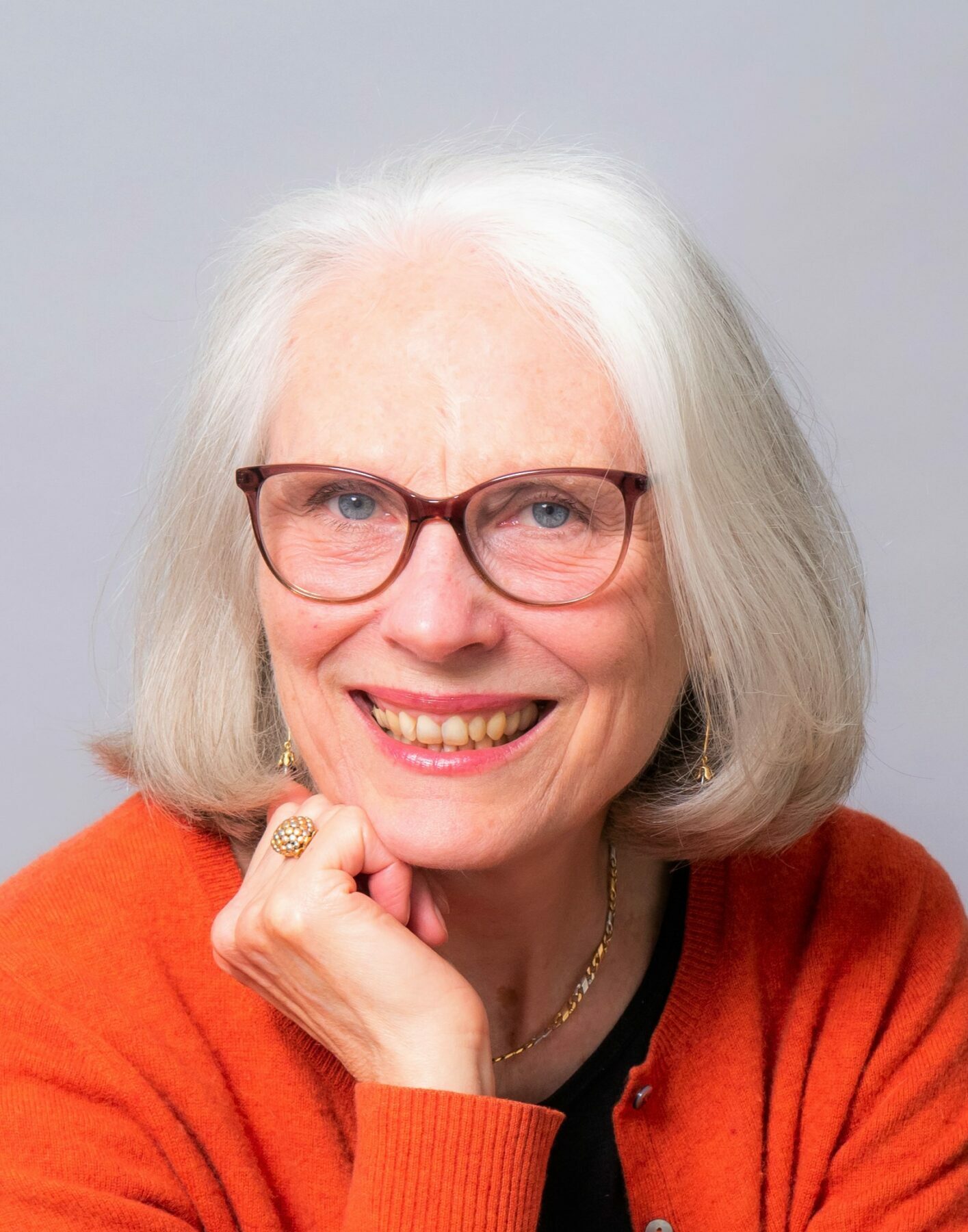 A woman with chin-length straight white hair and red rimmed glasses smiles, resting her chin on her fist