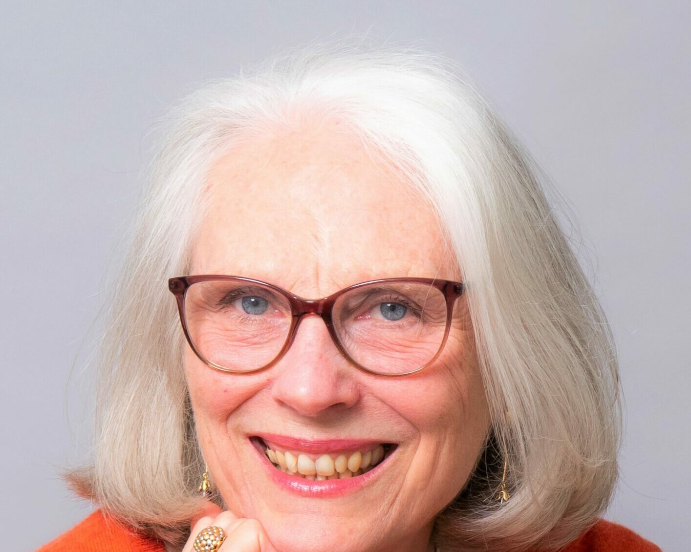 A woman with chin-length straight white hair and red rimmed glasses smiles, resting her chin on her fist