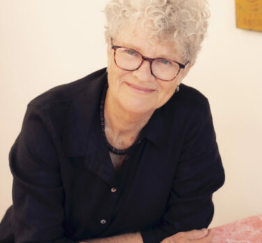 A woman with short curly gray hair and brown-rimmed glasses leans forward over a table, resting on her folded arms