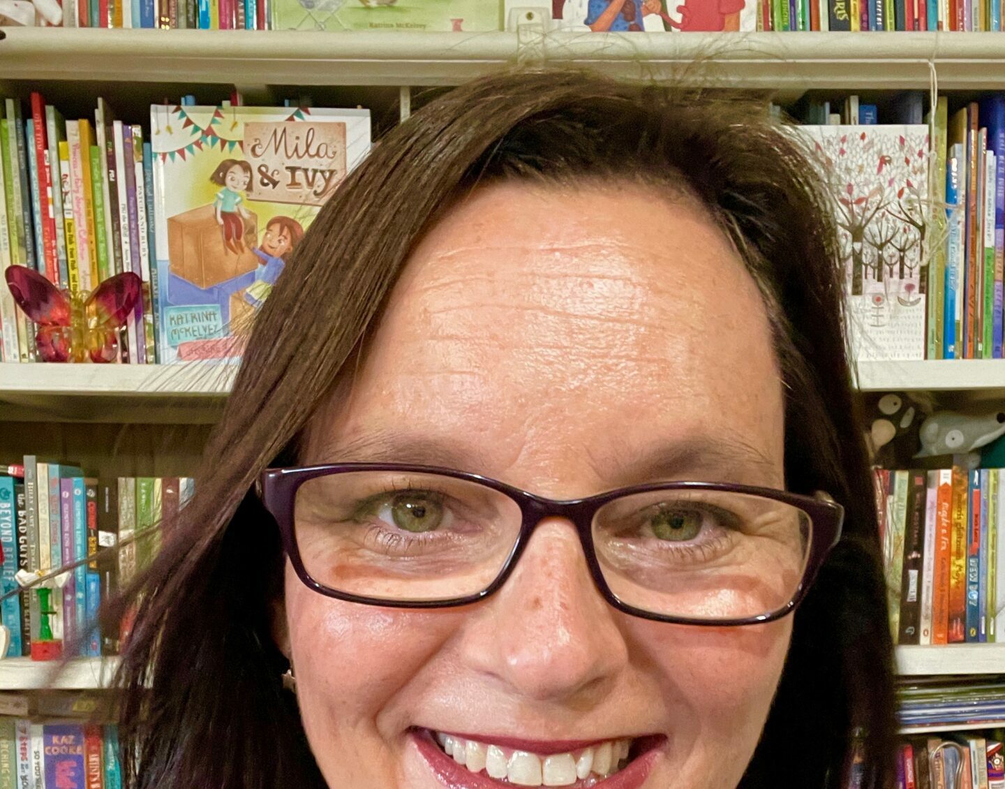 A close-up image of a white woman with long straight brown hair and glasses, smiling in front of a bookshelf